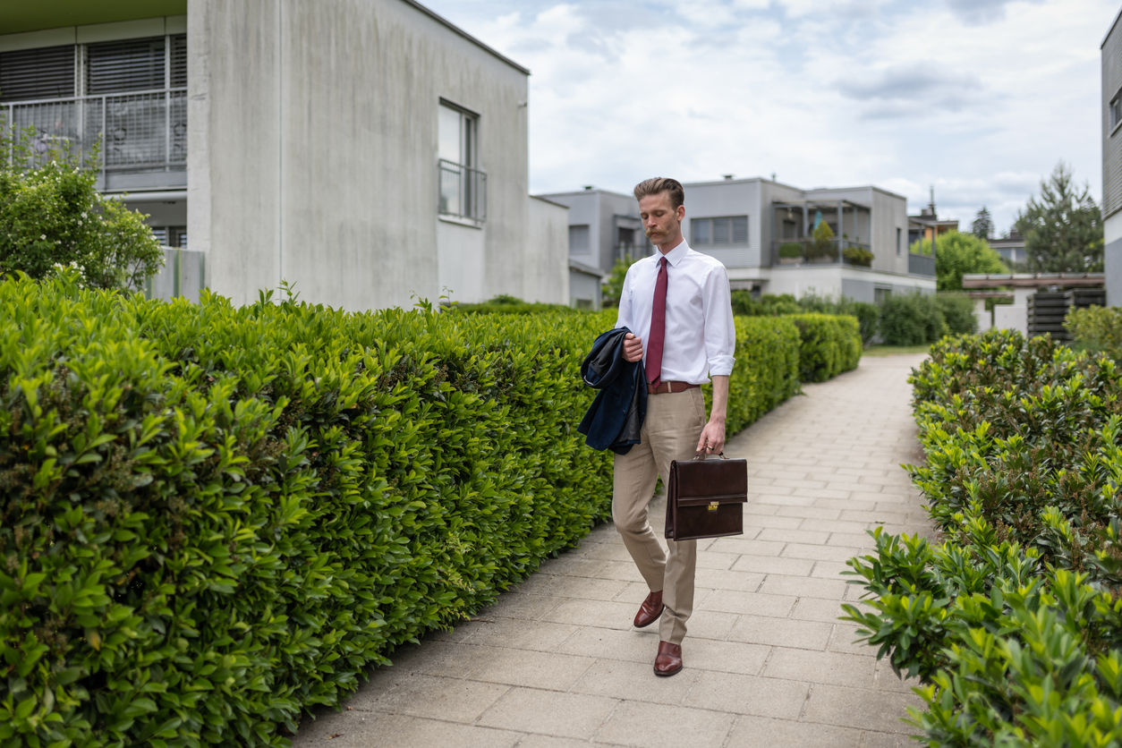 A man wearing a tie and holding a briefcase walks past gray condo buildings.
