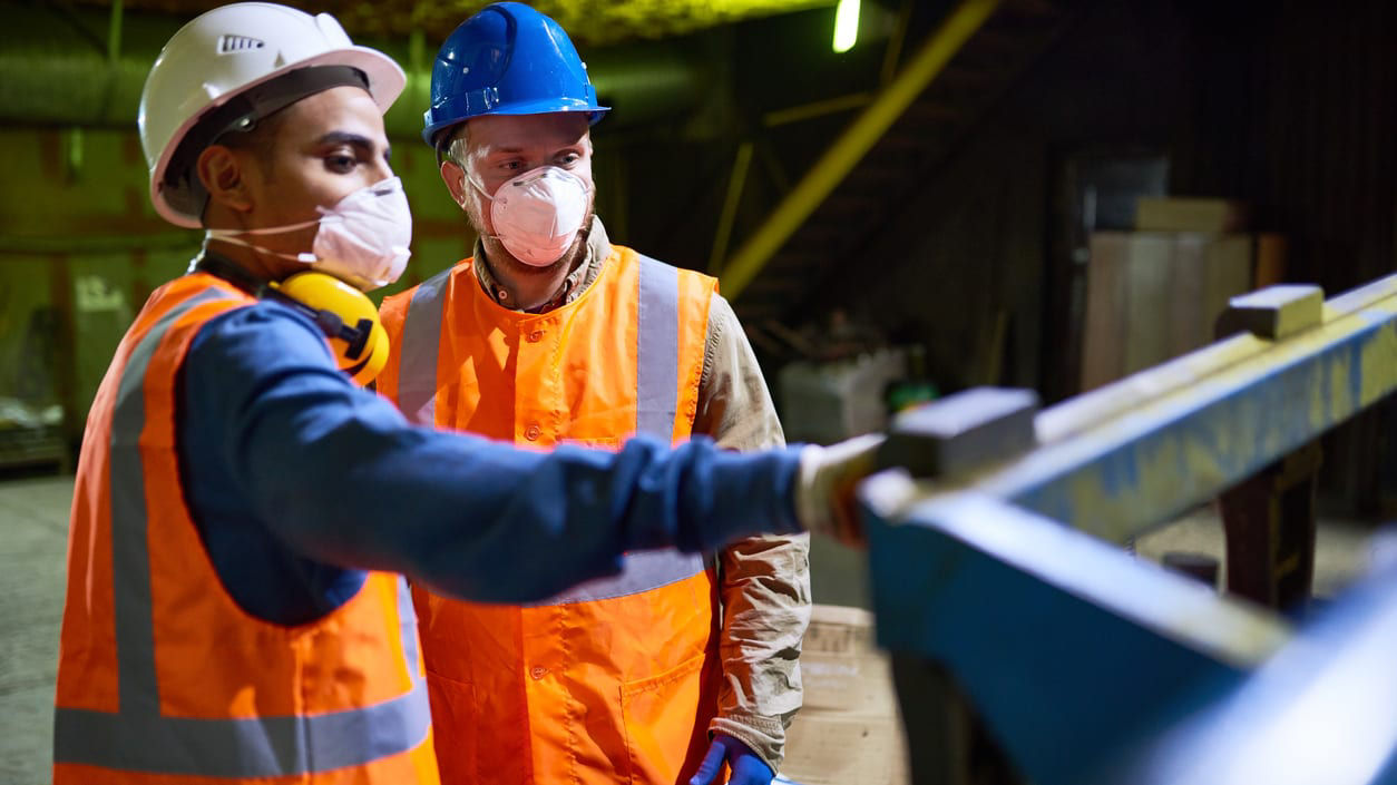 Two workers wearing safety vests and face masks in a factory.
