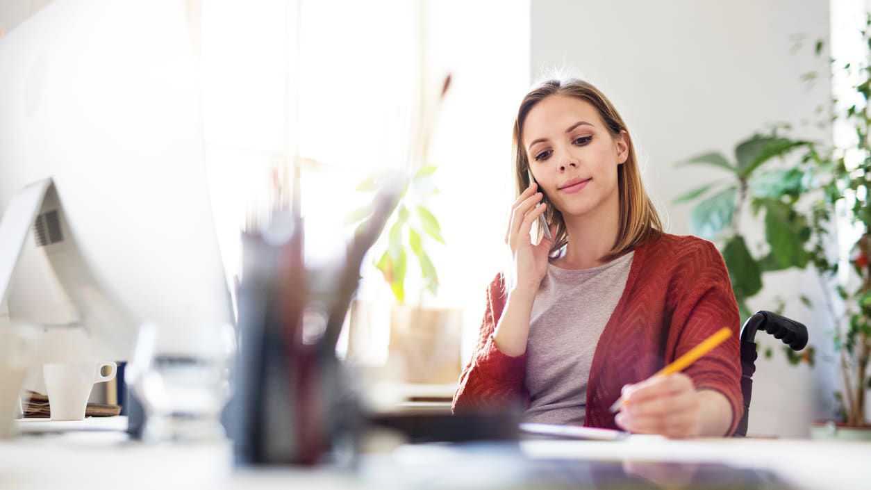 A woman in a wheelchair working at her desk with a pencil in her left hand to paper and her on a cellphone with her right hand.