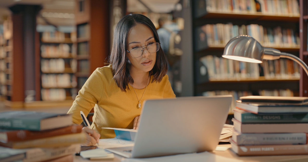 Person studying using their laptop. Young woman working on a case. Female alone inside a library working on a project