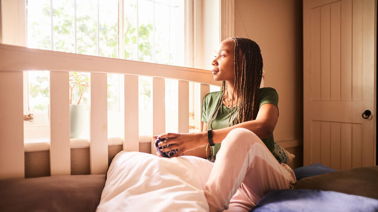 A young woman sitting on a bed looking out the window.