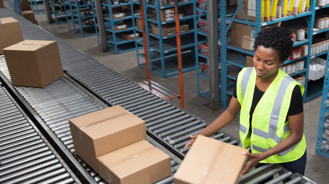 A woman is picking up boxes on a conveyor belt in a warehouse.