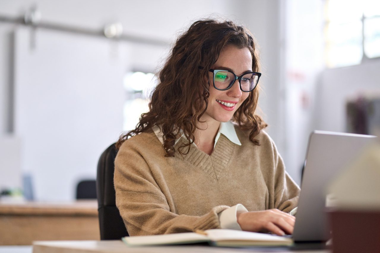 Young happy professional business woman worker employee sitting at desk working on laptop in corporate office. Smiling female student using computer technology learning online, doing web research.