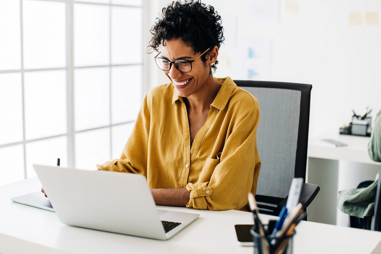 Graphic designer smiles as she works on a laptop in an office. Woman using a graphics tablet to make drawing designs. Creative business woman enjoys working on her project in an office.