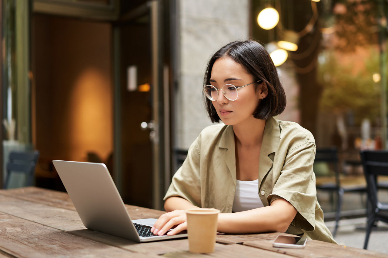 Young woman working in a cafe, using laptop and drinking coffee. Asian girl student with computer studying remotely, sitting on bench near shop.