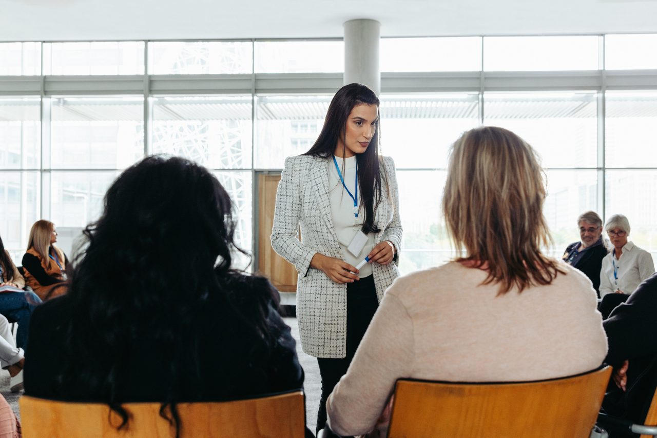 A small group meeting with people seated around the speaker, who is standing and listening.