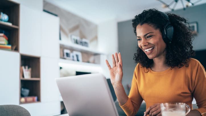 A woman wearing headphones is using a laptop and waving at the camera.