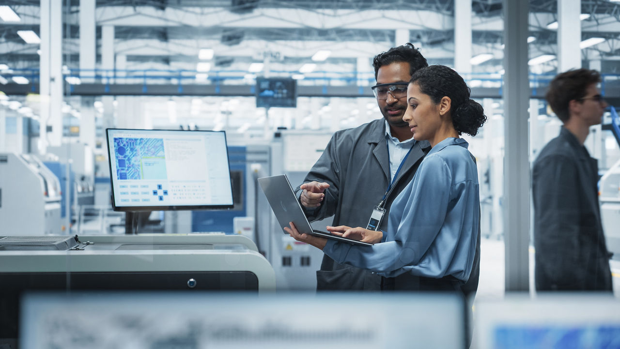 Indian Male Industrial Engineer And Hispanic Female Supervisor Using Laptop And Talking At An Electronics Factory. Man Using Soldering Jet Printer, Explaining Process Behind Production Of Motherboards