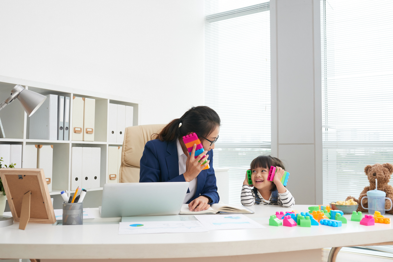 Adult Asian formal businesswoman with playful little girl sitting at table in modern office having fun with colorful blocks