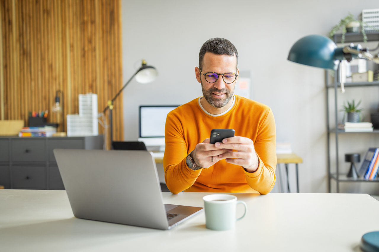 Businessman wearing casual clothing and eyeglasses sitting at his desk, using laptop and smart phone in the office.
