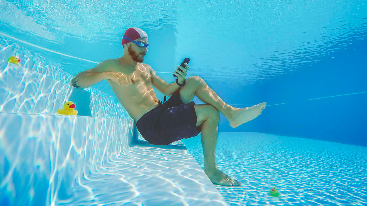 A swimmer holding a phone underwater with rubber ducks nearby.