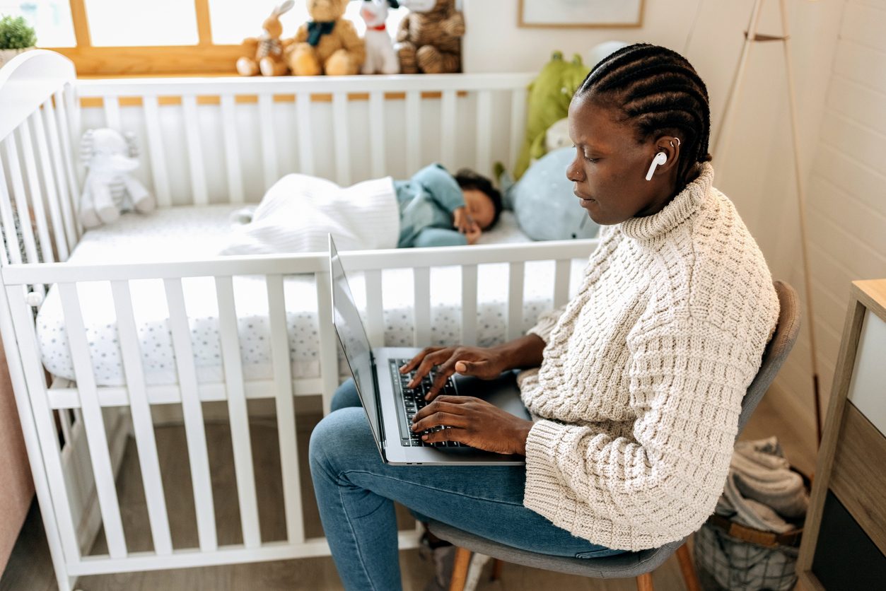 mother, sitting next to a crib, working on her laptop