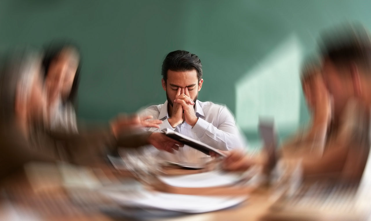 A man sitting with his fingers on the bridge of his nose with other people blurred around him.