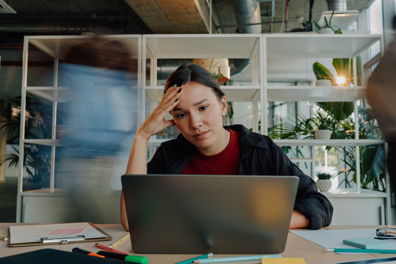 Young woman feeling overwhelmed and stressed while working intently on a laptop in a bustling office environment filled with activity