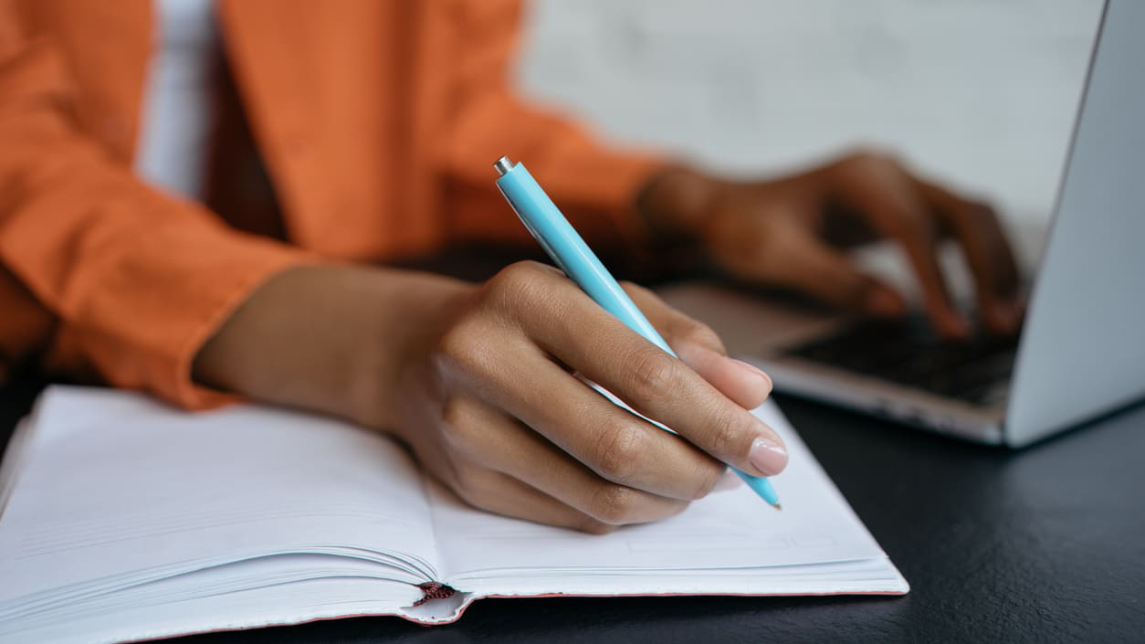 A woman's hand writing in a notebook next to a laptop.