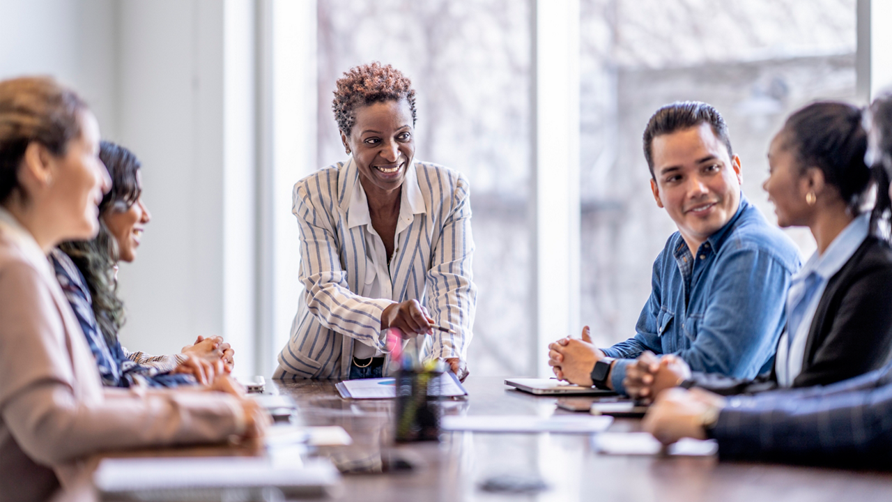 A small group of business professionals sit around a table as they meet to brainstorm some ideas for the future of the company. They are each dressed professionally and have papers scattered out in front of them as they work together. The focus is on a woman of African decent who is pitching an idea to the group.