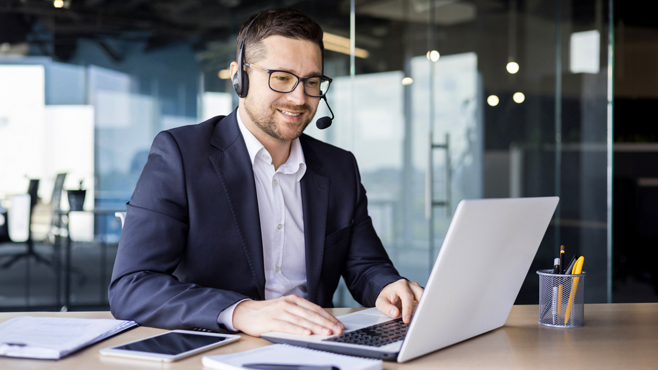 A young businessman is talking in the office on the news of a call from a laptop. He sits in the office in a headset, consults, supports, advises.