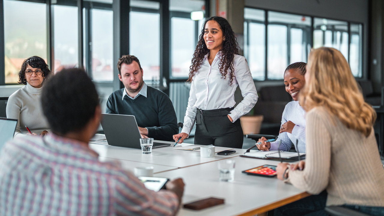Indoor shot of a big group of mixed race business people. They are seated around a table discussing a new project while taking notes on notebooks and laptops.