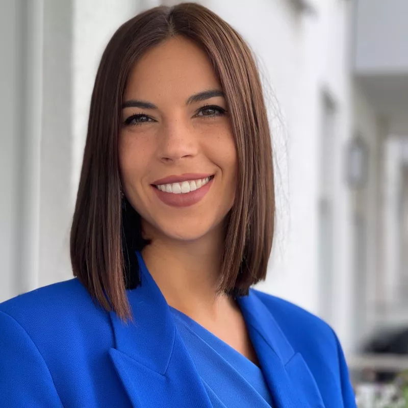 A professionally dressed woman stands outside wearing a bright blue blazer and matching top. Her straight, shoulder-length hair frames her blurred face, keeping her identity anonymous. The background shows a softly focused exterior corridor or building facade in neutral tones. The overall scene suggests a modern, corporate or business environment.