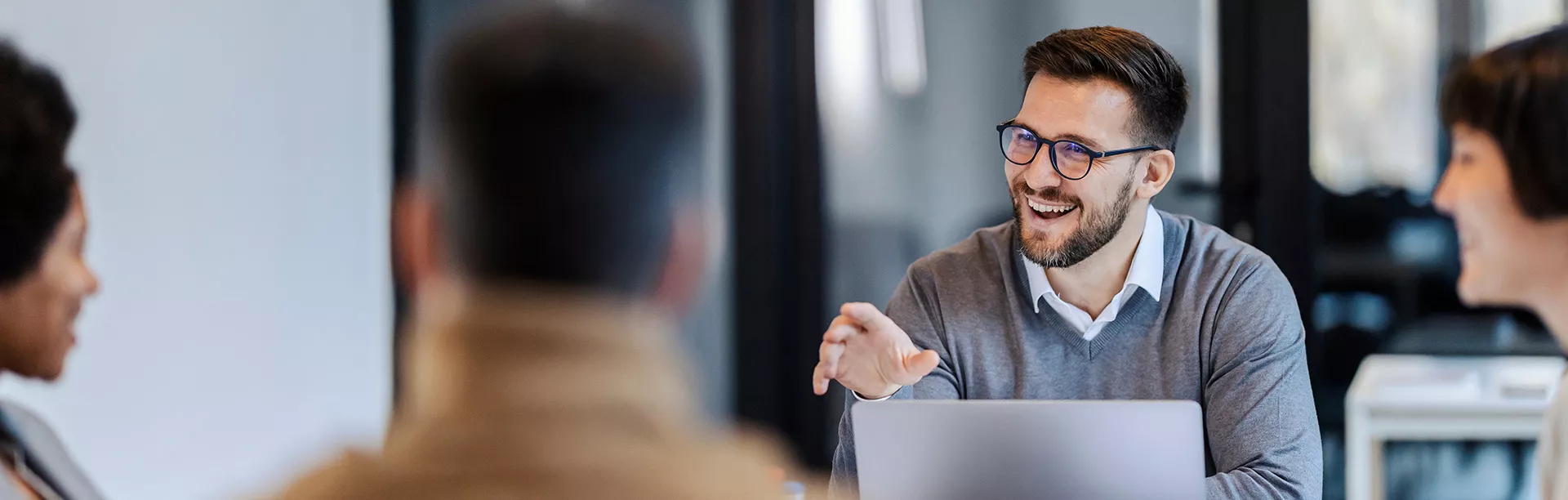 A group of professionals is engaged in a discussion around a table in a modern office setting. One person gestures while speaking, with a laptop open in front of them, suggesting a collaborative or brainstorming session. The environment features glass walls and a contemporary workspace design. The overall mood is focused and professional, with neutral tones dominating the palette.