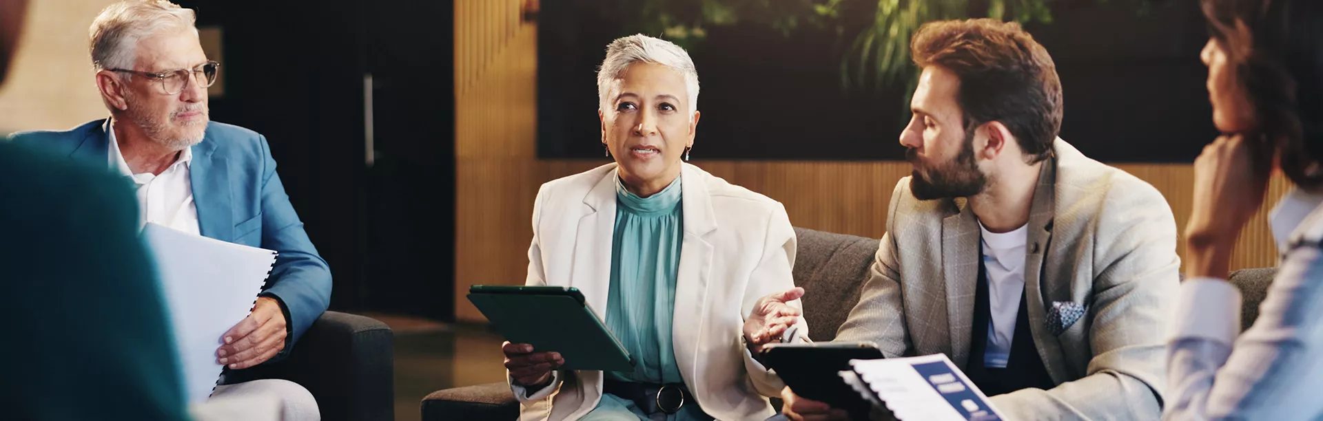 A group of business professionals are engaged in a discussion, seated together in a modern office environment. One person holds a tablet, while another has documents, suggesting a collaborative or brainstorming session. The setting features contemporary decor and a relaxed yet focused atmosphere. The group includes both men and women of varying ages, dressed in business attire.
