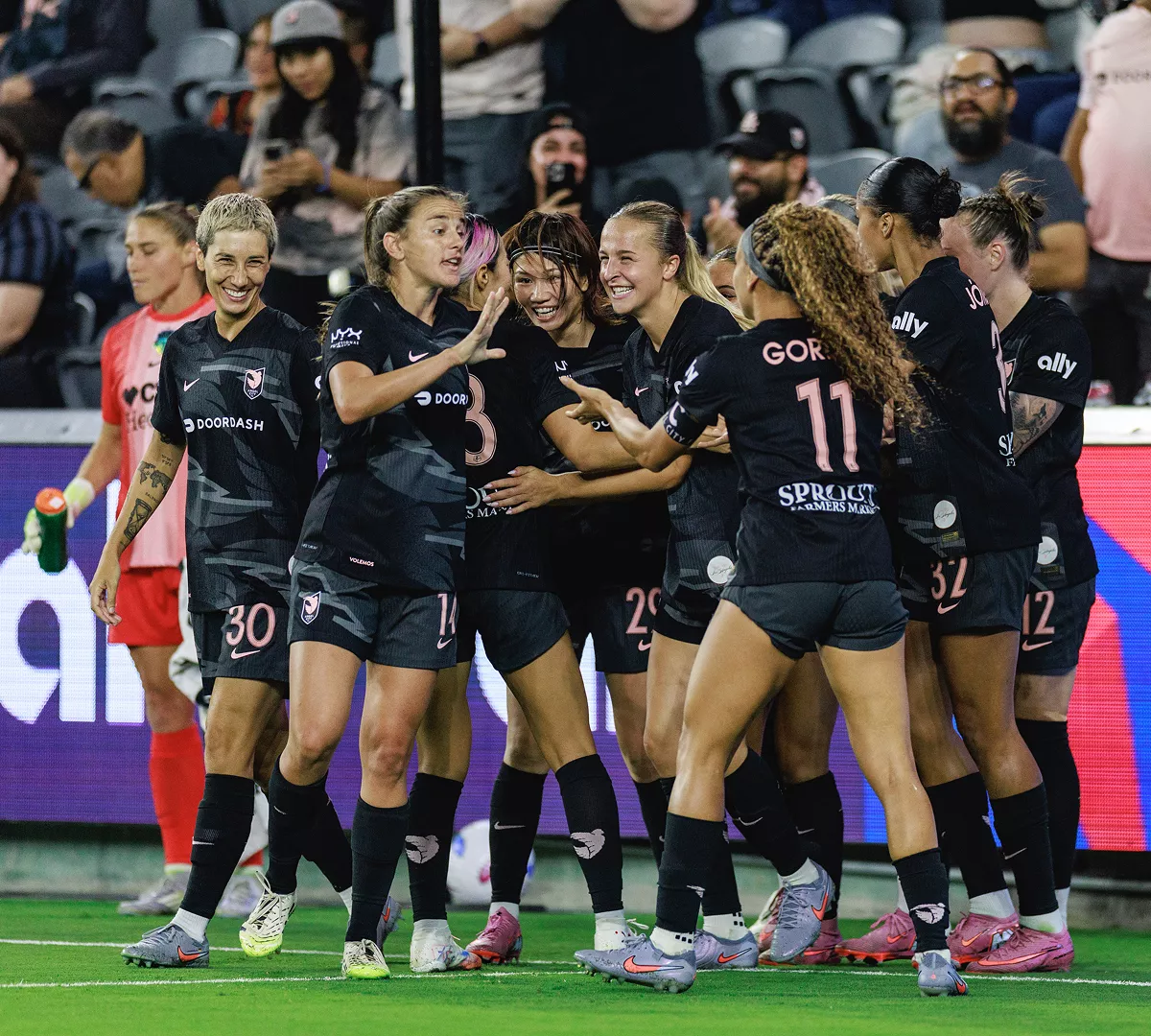 A women’s soccer team gathers in a tight huddle on the field, celebrating during a match. The players wear dark uniforms with visible numbers including 30 and 11 on their jerseys. Stadium seating and spectators fill the background, suggesting a professional or competitive environment. Bright field lighting and vivid colors emphasize the energy and excitement of the moment.