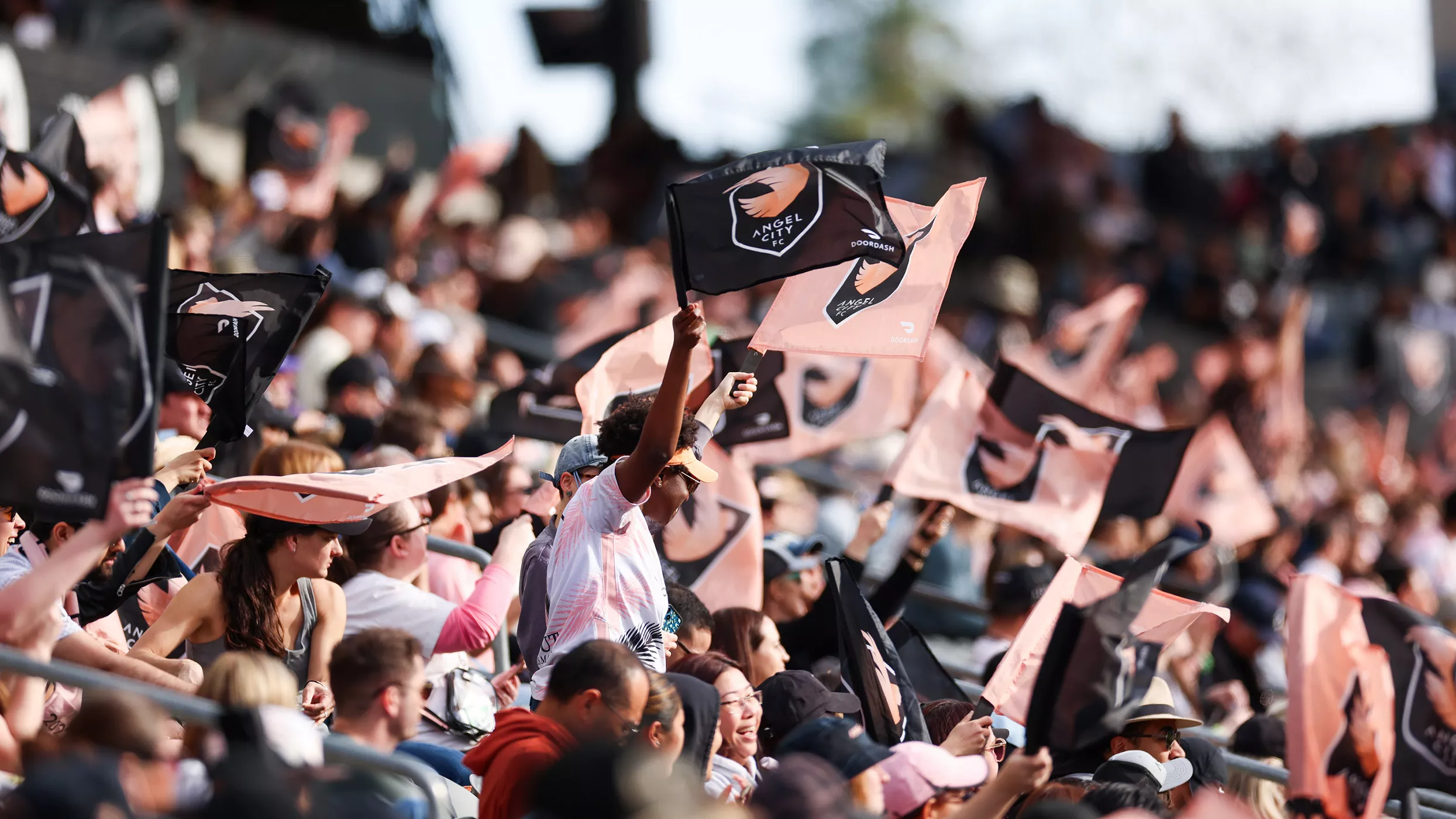 A large crowd of fans fills the stadium stands, enthusiastically waving matching team flags. The scene captures the energy of a live sporting event with people standing, cheering, and raising their arms. Black and peach-colored flags dominate the view, creating a dynamic pattern across the audience. Bright daylight and shallow depth of field emphasize the excitement and movement in the crowd.