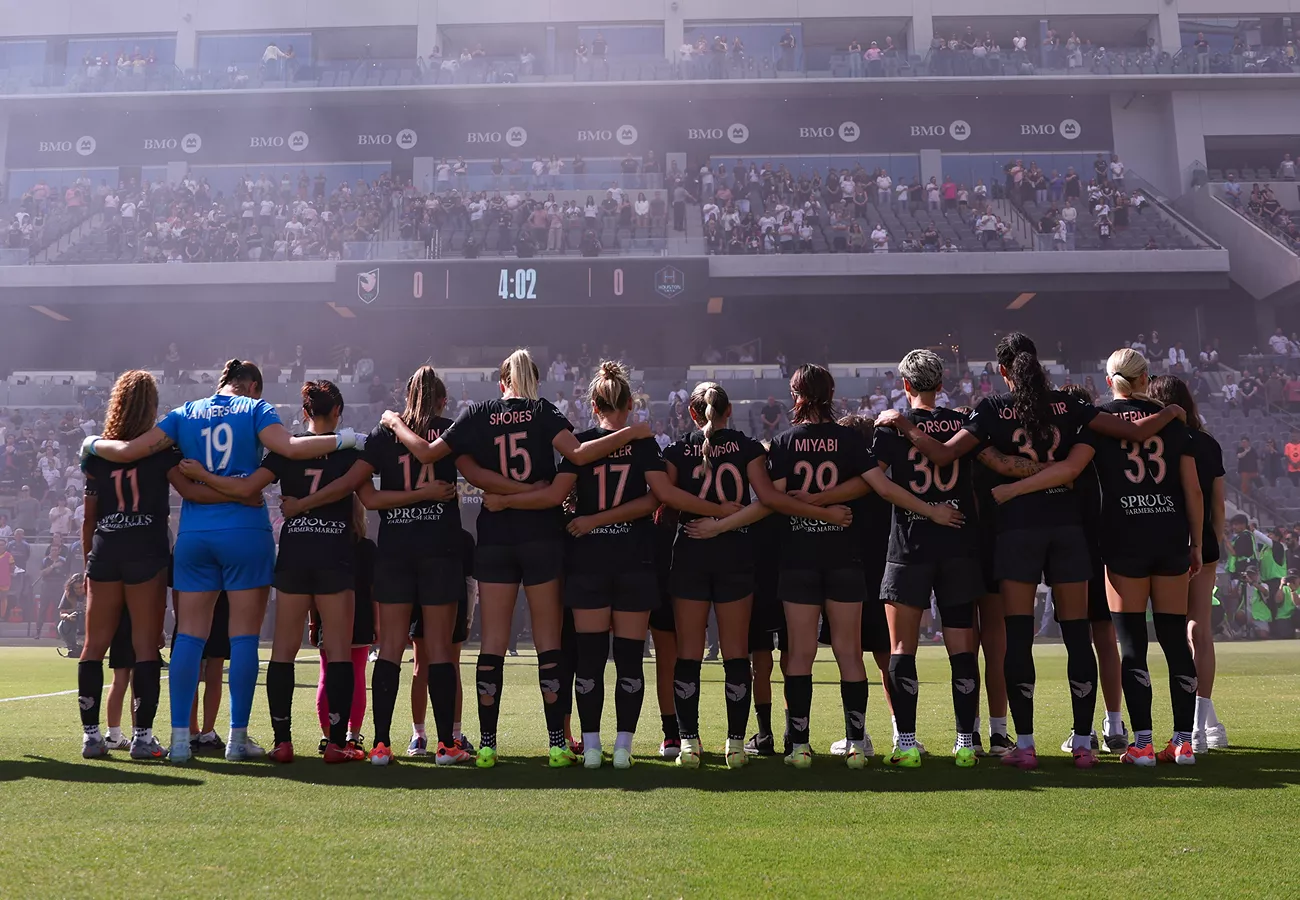 A women’s soccer team stands arm in arm on the field facing a packed stadium. The players wear dark uniforms with visible jersey numbers including 19, 15, 17, 10, 23, and 26. Sunlight and light haze create a dramatic atmosphere as the crowd watches from the stands. The scene captures unity, anticipation, and team spirit before kickoff.
