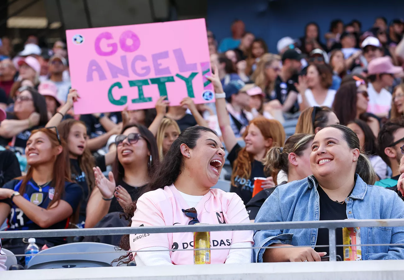 A crowd of spectators sits in a stadium, with one fan prominently holding a bright pink sign that reads "GO ANGEL CITY." The audience is dressed casually, many wearing dark shirts and sports apparel, suggesting support for a team. Faces are blurred, but the scene conveys excitement and community. Metal railings and drink bottles are visible in the foreground, emphasizing the live event atmosphere.