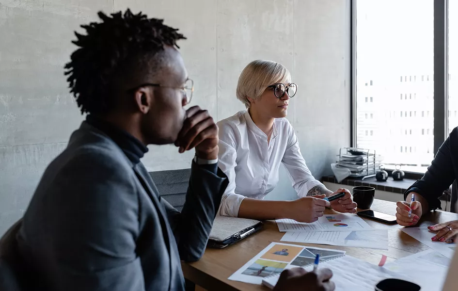 A group of professionals is gathered around a conference table in a modern office setting. The table is covered with printed charts, graphs, and reports, indicating a business discussion or strategy session. Natural light streams in from a large window, and the mood is focused and collaborative. Visible items include pens, coffee mugs, and electronic devices.
