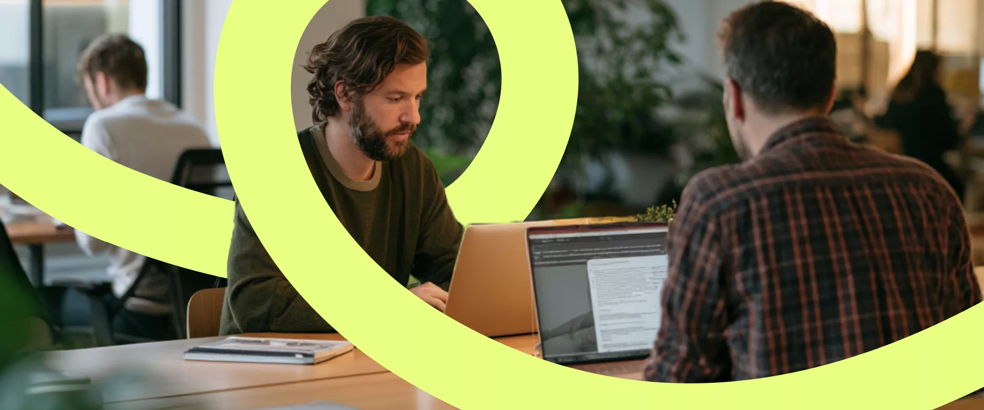 Two men work on laptops at a shared desk in a contemporary open-plan office. One faces the camera while concentrating on his screen, and the other has his back turned, reading from his laptop. Additional coworkers are visible in the background, creating a busy yet focused atmosphere. Bright, curved neon-yellow graphic elements overlay the scene, adding a dynamic and creative visual accent.