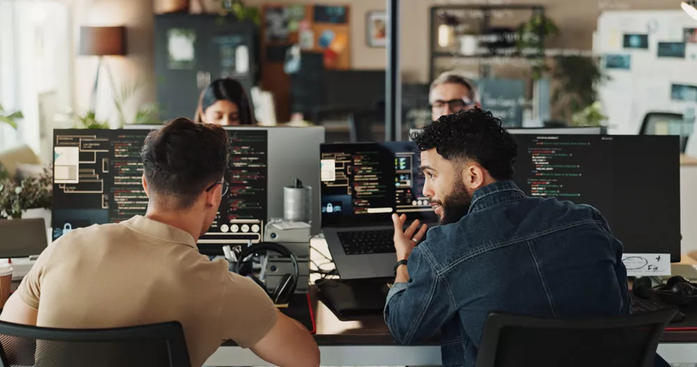 The image shows two young adults working together at a desk with multiple computer monitors displaying code. The setting is a contemporary office with a casual atmosphere, featuring blurred background elements such as shelves and plants. The workspace is well-lit, and the mood is focused and collaborative, with neutral and earthy tones dominating the palette.
