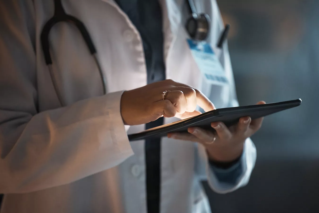 A medical professional in a white coat interacts with a digital tablet, likely reviewing patient information or medical records. The setting appears to be a healthcare environment, indicated by the stethoscope and identification badge. The image is softly lit, focusing on the hands and device, with no visible text or numbers. The mood is professional and modern, emphasizing technology in medicine.