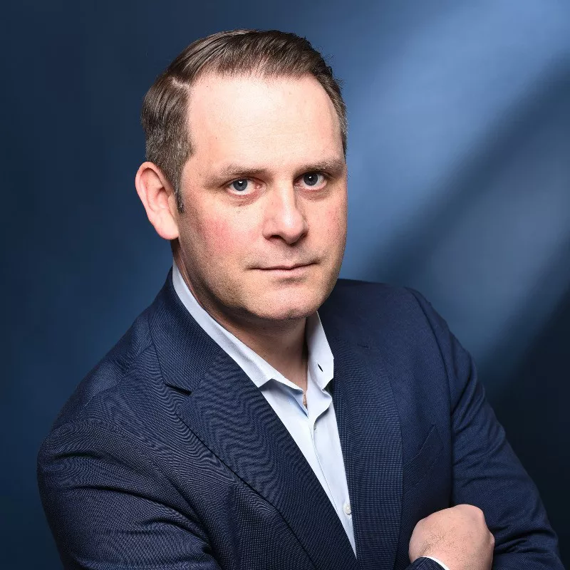 A person is photographed in a studio setting wearing a navy blue suit and white dress shirt. The background features a gradient blue tone with soft lighting, creating a formal and polished atmosphere. The subject's face is obscured, and no visible text or numbers are present in the image.