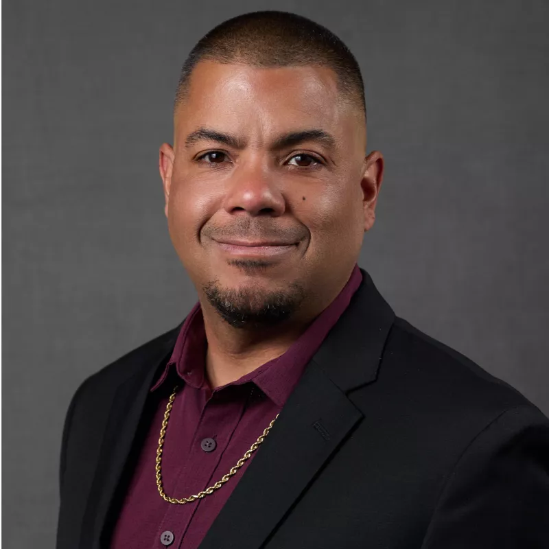 A man poses for a formal studio portrait against a plain gray background. He is dressed in a dark blazer over a burgundy shirt, accessorized with a gold chain necklace. The subject looks directly at the camera with a slight smile, conveying confidence and approachability. The lighting is even and soft, highlighting his face and professional appearance.