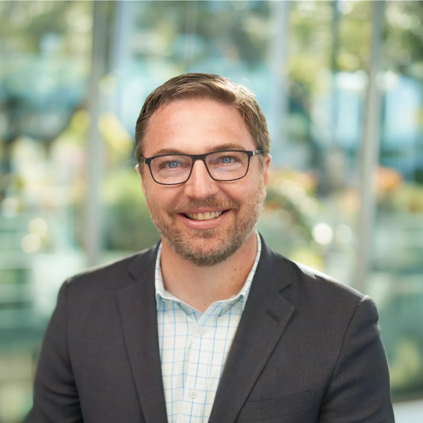A smiling adult man wearing glasses and a suit jacket poses for a portrait. He stands in front of large glass windows with a bright, blurred office or corporate environment behind him. Soft natural light highlights his face and creates a warm, approachable mood. The overall scene suggests a professional business setting or corporate headshot.