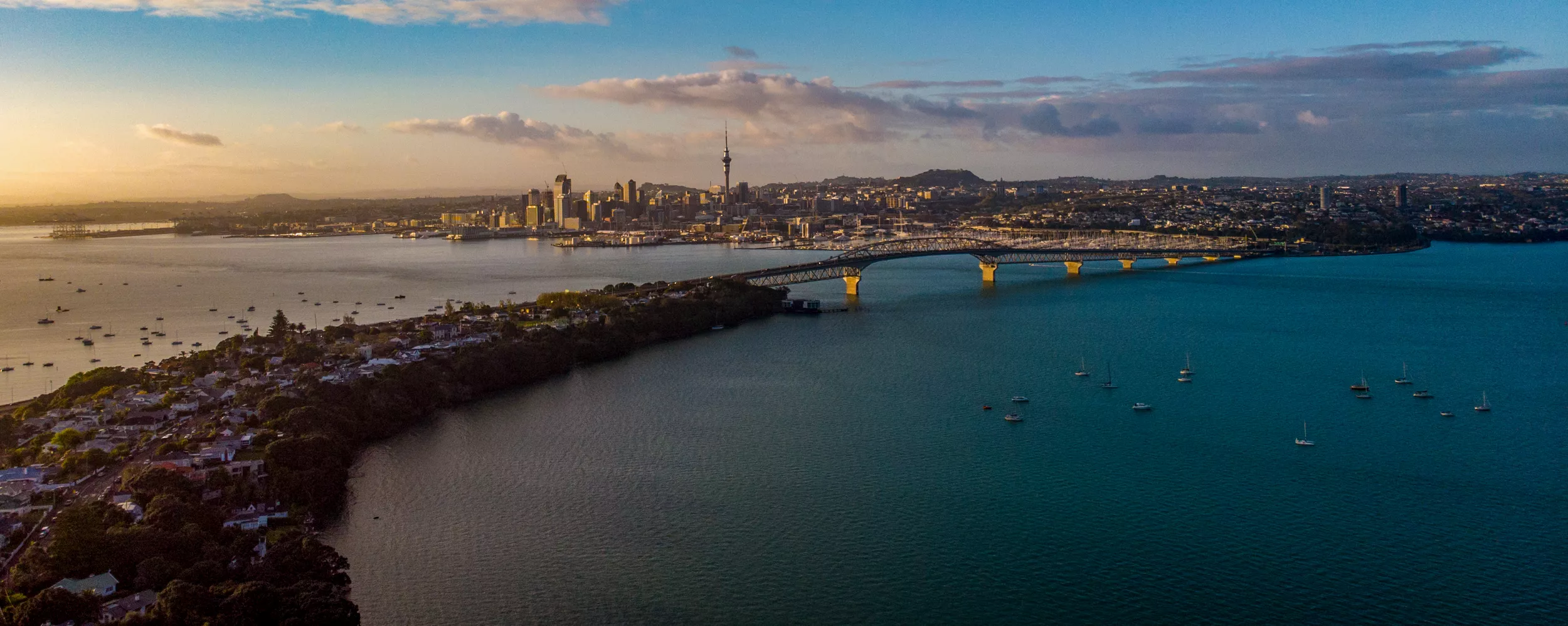 A wide aerial panorama shows a coastal city skyline at sunset, with warm light illuminating the buildings in the distance. A long bridge spans the water, connecting the urban center to a greener residential shoreline in the foreground. Numerous small boats are scattered across the calm harbor, reflecting the soft evening colors. The sky features gentle clouds transitioning from golden hues near the horizon to cooler blues overhead.