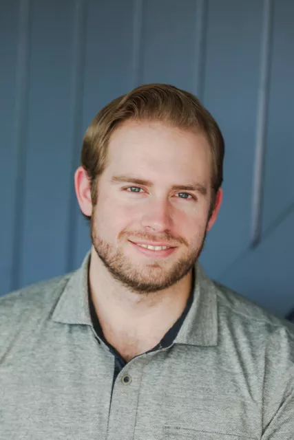 Portrait of a man smiling softly at the camera. He is wearing a casual grey polo shirt and has neatly styled short hair and a trimmed beard. The background features vertical blue panels, creating a clean and modern setting. Soft natural lighting highlights his face, giving the image a friendly and approachable feel.