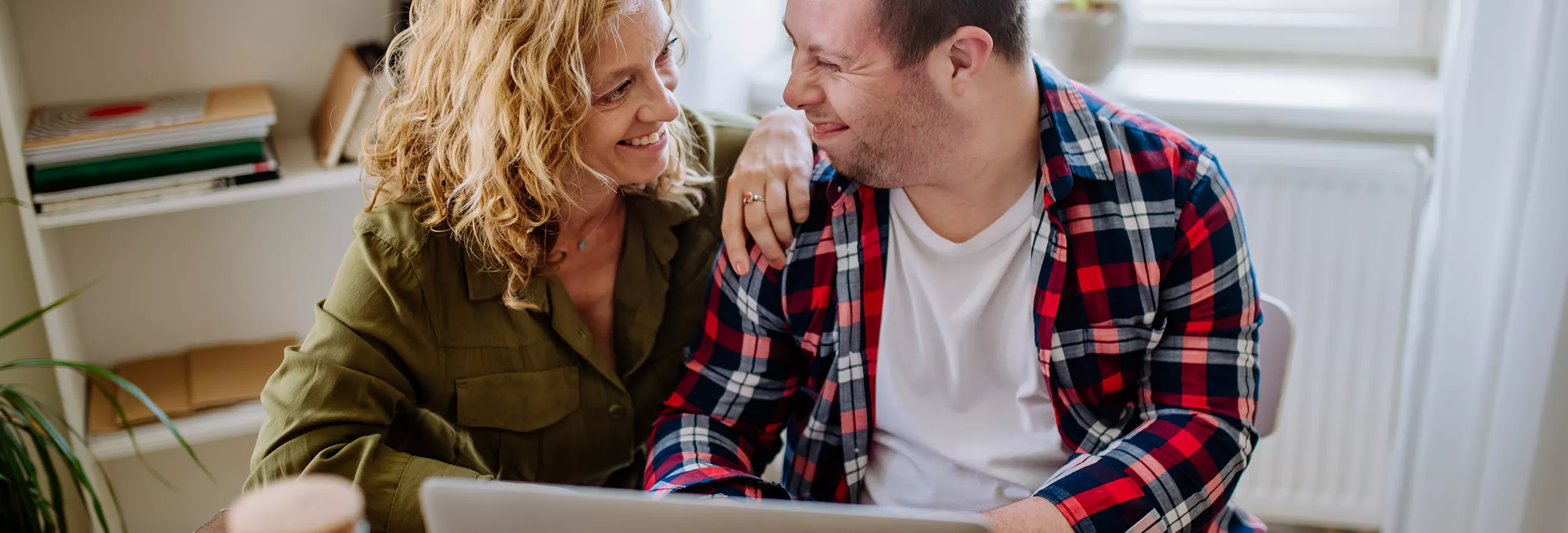 A man and woman sit closely together at a table, smiling warmly at each other while using a laptop. The scene appears to be in a bright home interior with a window in the background. The man wears a red and blue plaid shirt, and the woman wears an olive green top. Their relaxed posture and eye contact suggest a supportive, affectionate relationship.