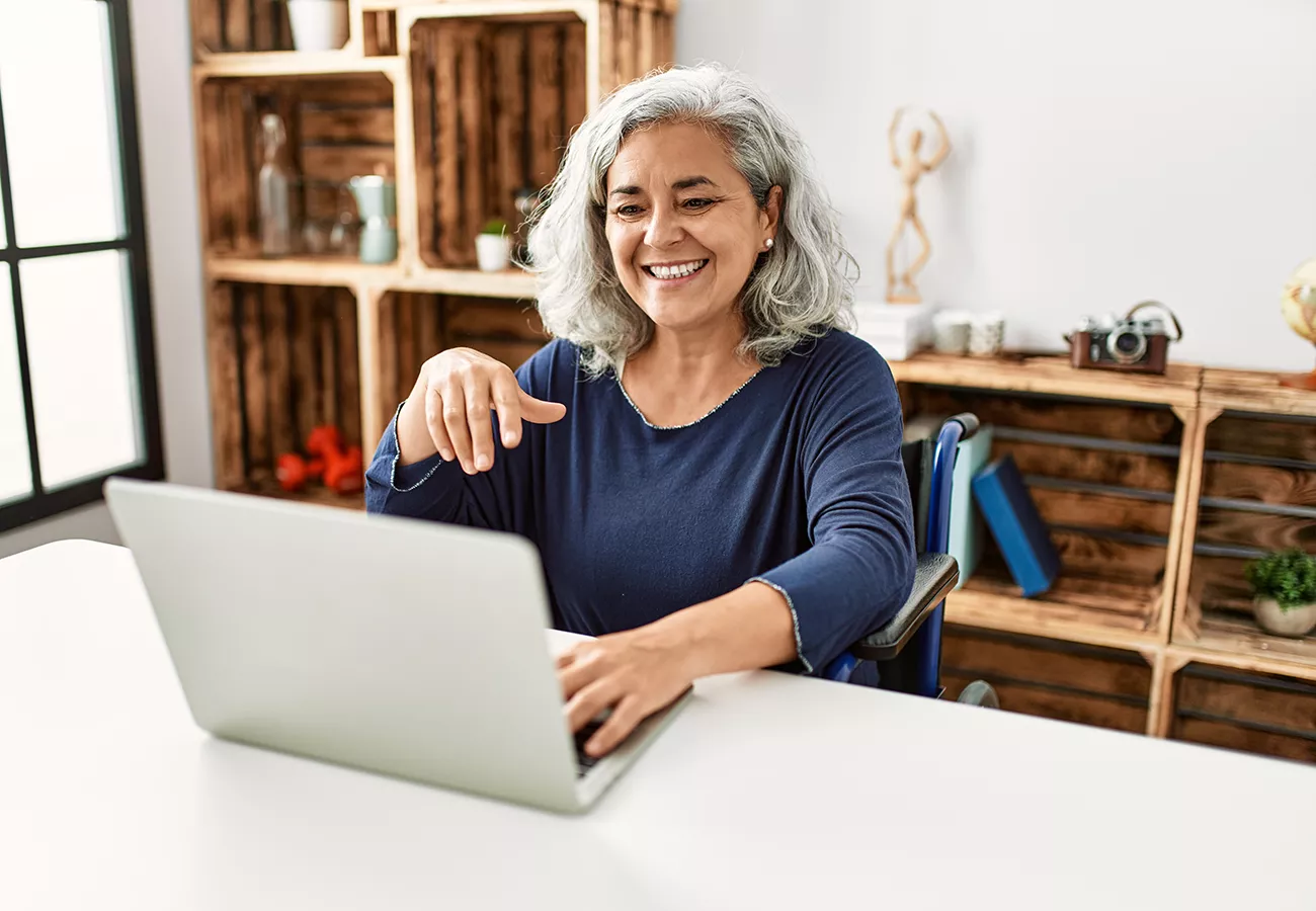 An older woman with gray hair sits at a desk using a laptop, smiling and gesturing toward the screen. She appears to be in a cozy home office with wooden shelves and decorative items in the background. Natural light comes from a large window, creating a warm and inviting atmosphere. The scene suggests remote work, online communication, or casual browsing in a comfortable setting.