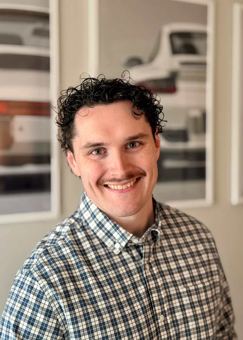 A man wearing a blue and white checkered button-up shirt stands indoors. The background features framed photographs or prints of classic cars, adding a modern and stylish touch to the setting. The overall mood is casual and professional, with neutral tones and soft lighting. No visible text or numbers are present in the image.