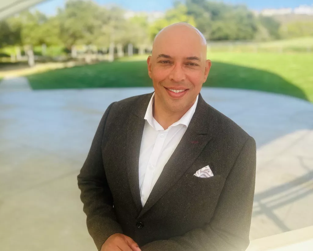 A bald man in a dark suit jacket and white dress shirt smiles at the camera. He stands under a light-colored structure with a concrete walkway behind him. A large green lawn and trees stretch out into the background under bright daylight. The overall scene feels professional yet relaxed in an outdoor setting.