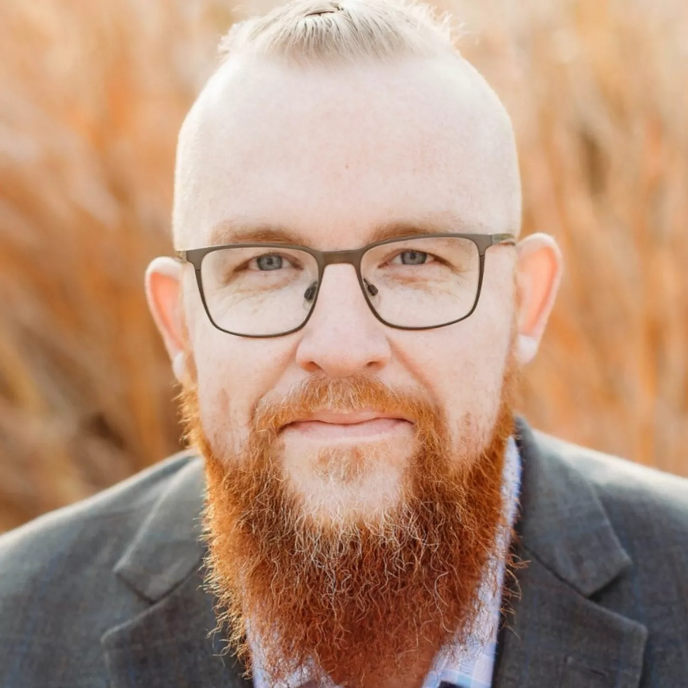 Close-up portrait of an adult man with a long reddish beard and glasses. He is looking directly at the camera with a neutral, slightly warm expression. The background is softly blurred, featuring golden-brown outdoor foliage that suggests a natural setting. Warm lighting enhances the earthy tones and gives the image a calm, approachable feel.