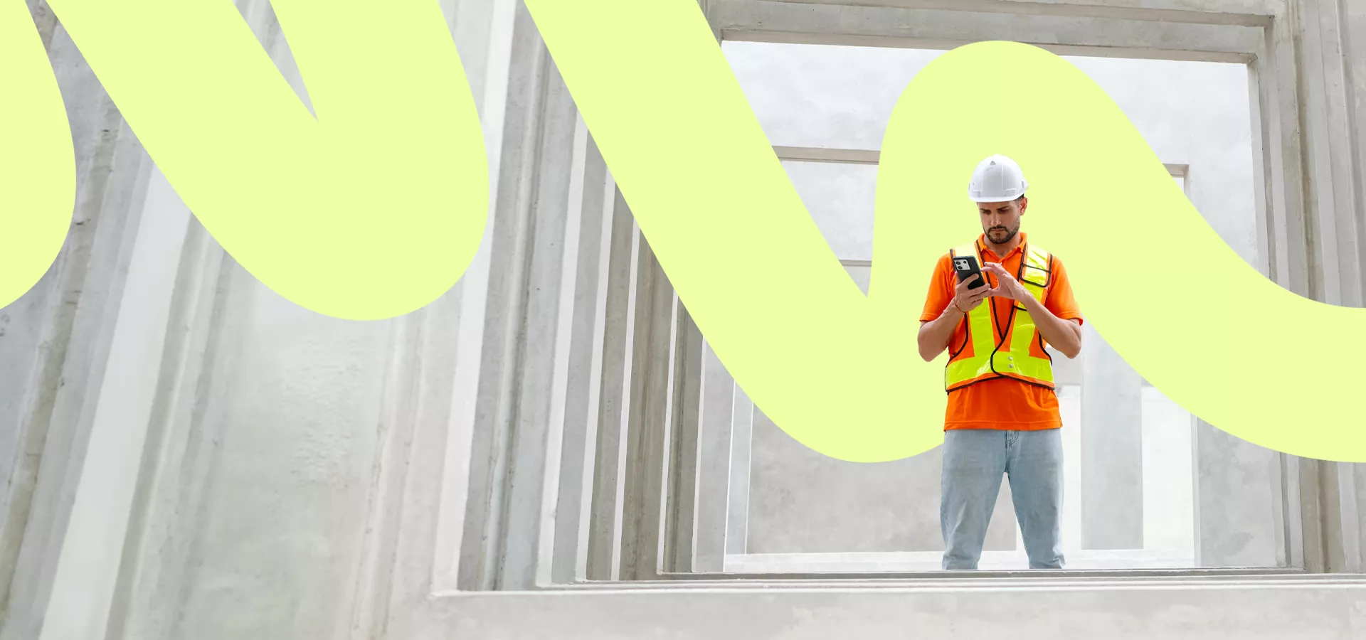 A construction worker wearing a white hard hat and orange safety vest stands between large concrete structures. He is focused on a handheld mobile device, suggesting digital tools or inspection work on the job site. The architectural setting features repeating rectangular frames and industrial materials. A bold, light yellow graphic wave overlays the scene, adding a modern, creative visual element.