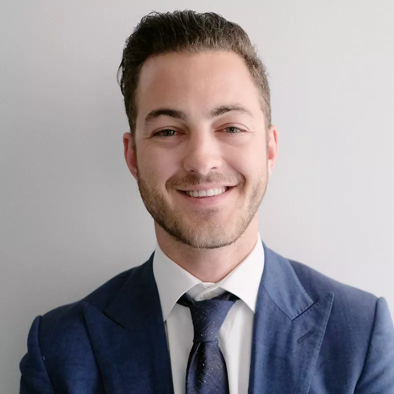 A young adult man is shown in a close-up portrait wearing a blue suit, white dress shirt, and dark tie. He is smiling warmly while standing against a plain light-colored wall. The image has soft, even lighting that highlights his face and professional appearance. The overall style is clean, modern, and corporate, suitable for business or professional profile use.