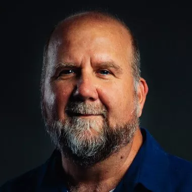 A mature man with a bald head and gray beard is photographed against a dark background. He is wearing a dark blue collared shirt, and the lighting highlights his facial features and upper torso. The overall mood is serious and subdued, with a focus on the subject's expression and attire. No visible text or numbers are present in the image.