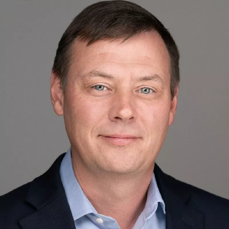 A man dressed in a dark suit jacket and light blue collared shirt is pictured against a plain gray backdrop. The subject appears to be an adult male, with short brown hair, and is shown from the shoulders up. The setting is neutral and professional, suitable for corporate or business contexts. No visible text, logos, or numbers are present in the image.
