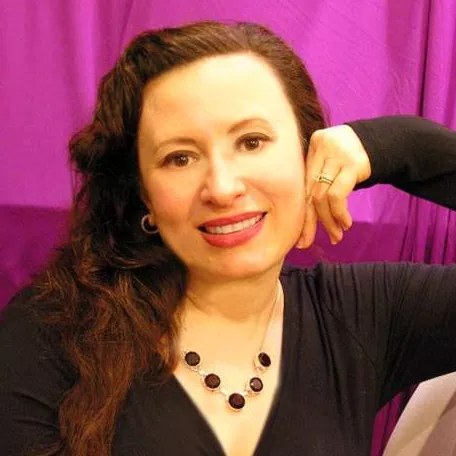 A woman with long dark hair smiles at the camera while resting her head on her hand. She wears a dark top and a beaded necklace, along with a ring on her finger. The background features rich purple fabric that creates a warm, studio-like setting. The overall mood is friendly and approachable, with soft lighting highlighting her face.