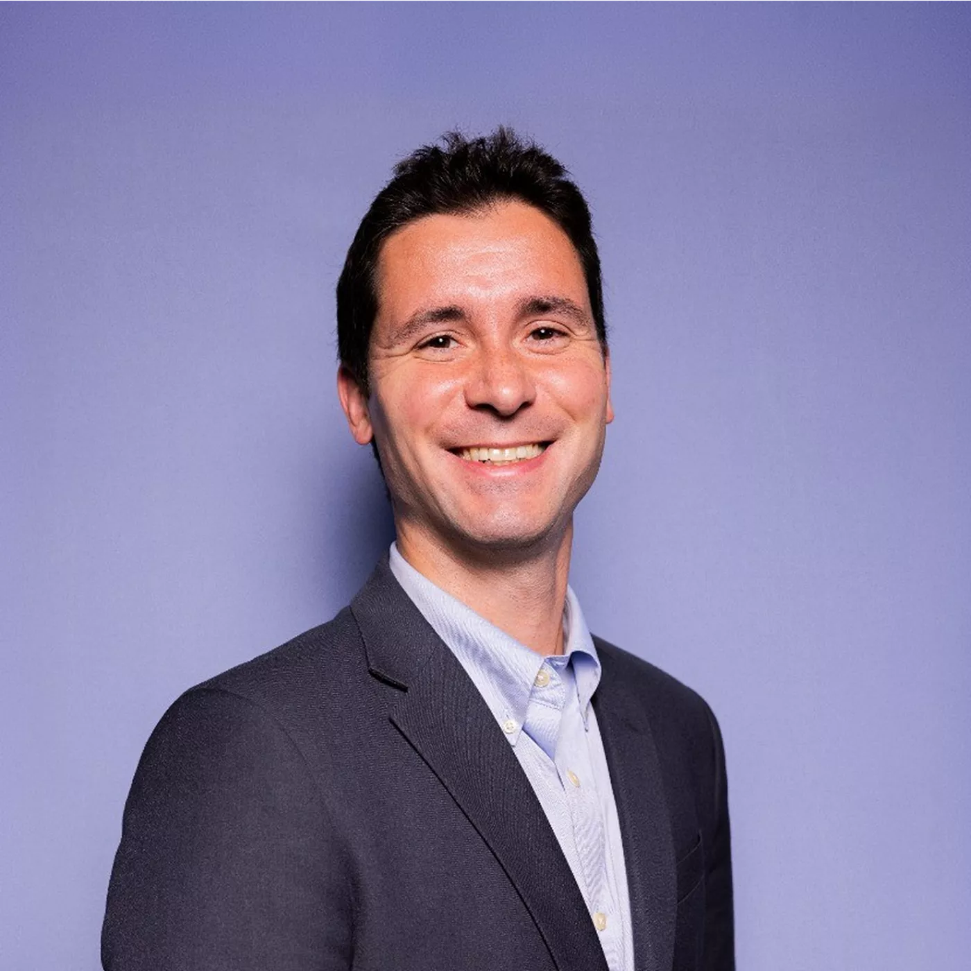 A man in business attire smiles confidently while posing for a professional portrait. He is wearing a dark blazer over a light button-up shirt. The subject stands against a solid light purple background, creating a clean and modern studio look. The image conveys approachability and professionalism, suitable for corporate or business-related uses.