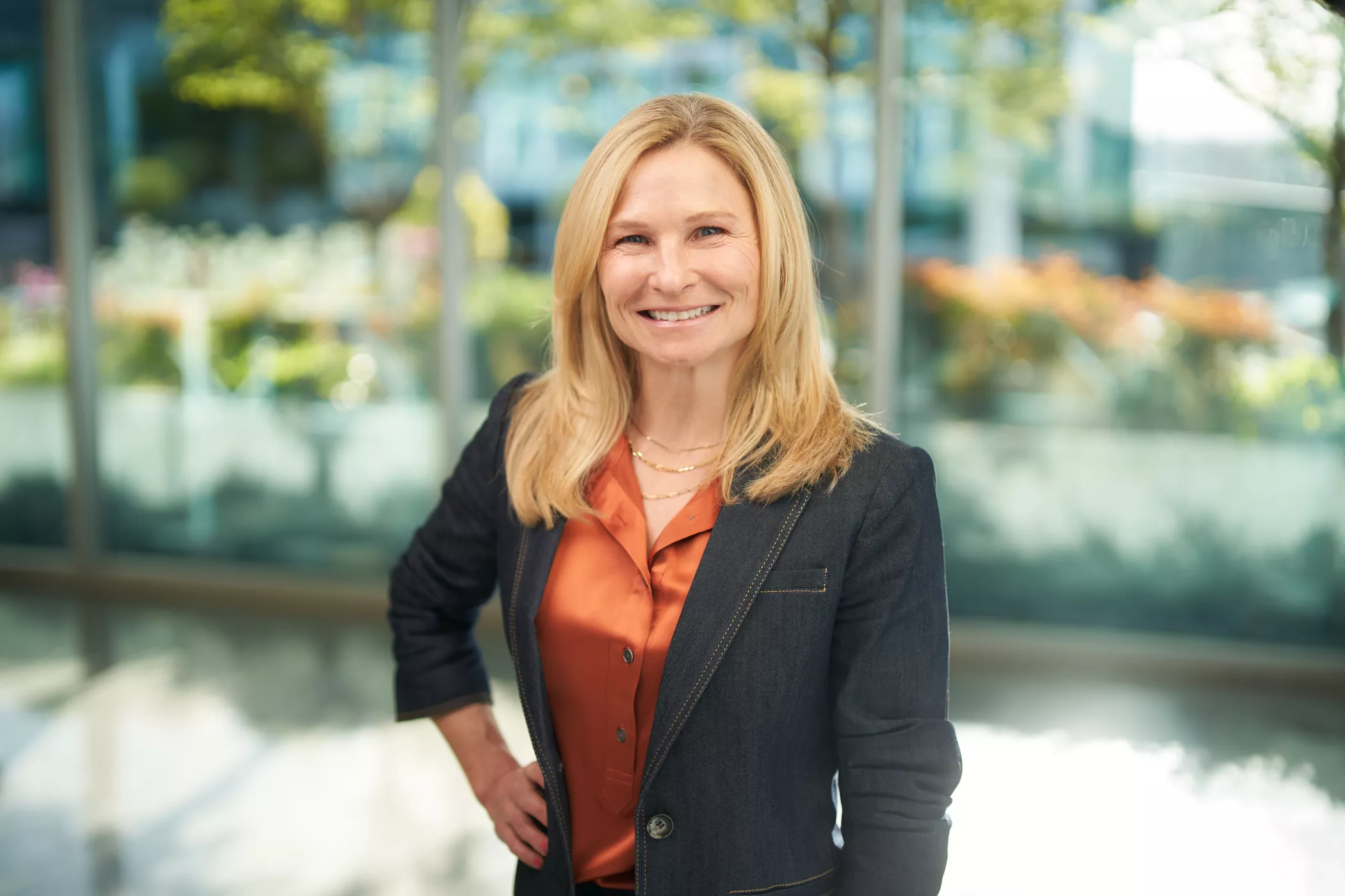 A blonde-haired woman wearing a dark blazer and orange blouse stands confidently in a bright, modern office environment. The background features large glass windows with a view of greenery and flowers. The setting suggests a professional or corporate atmosphere.
